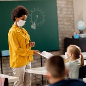 Happy black teacher giving exam paper to her student while wearing protective face mask in the classroom. Happy black teacher and her students wearing protective face mask in the classroom. Teacher is giving them their test results. (Happy black teacher and her students wearing protective face mask in the classroom. Teacher is giving them their test resu